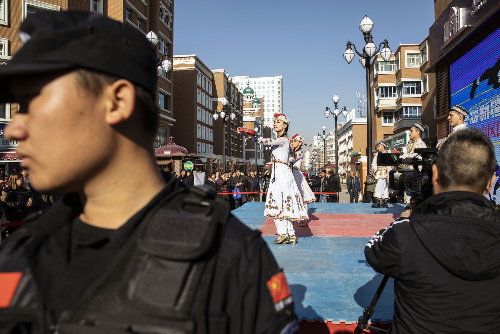 Một nhân viên an ninh đang làm việc trong khi những phụ nữ đang biểu diễn điệu múa cổ truyền ở khu chợ chính trong thủ đô Ô Lỗ Mộc Tề của Tân Cương hồi Tháng Mười Một ( có lẽ là 2018 -- chú thích của người dịch. ) Nguồn: Bloomberg